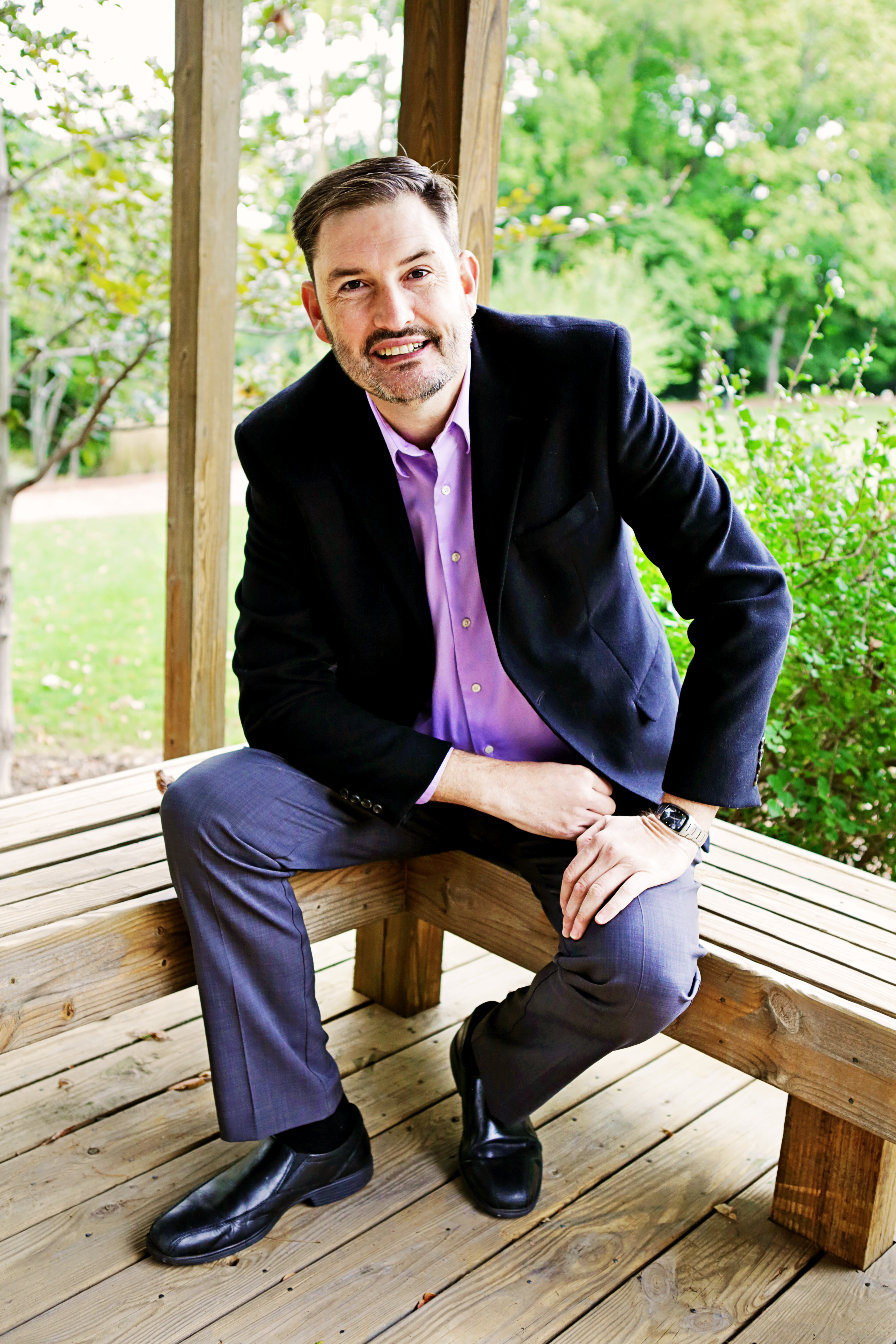 Michael Harshbarger seated on a wooden bench outdoors, wearing a dark blazer and lavender shirt and smiling toward the camera.